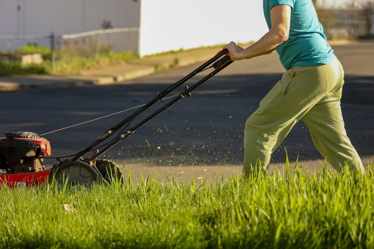 Man pushing a red lawn mower, cutting tall green grass on a sunny day